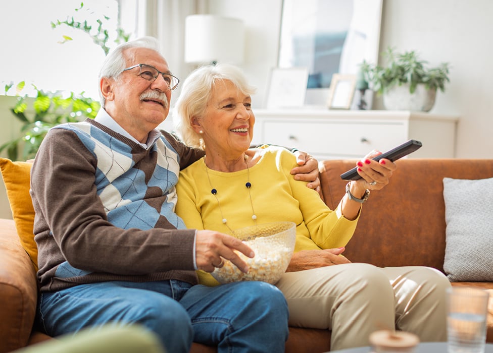 senior man and woman sitting on the couch enjoying popcorn while watching a movie